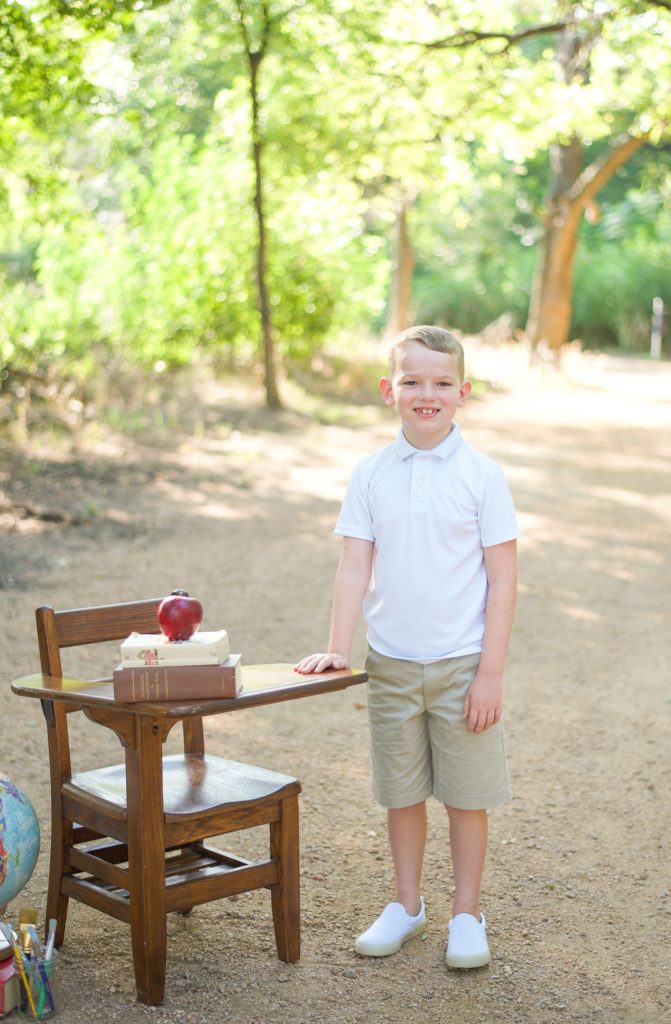 boy stands by wooden desk with apple on books for Back to School Mini Sessions