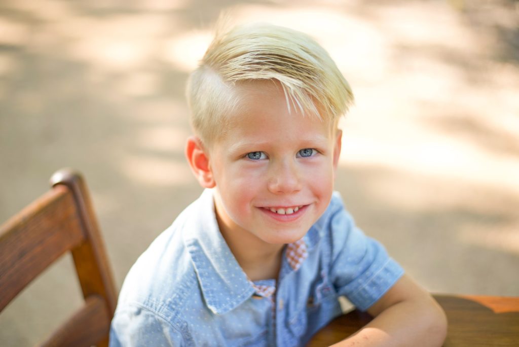 young boy in blue shirt leans on wooden desk and smiles