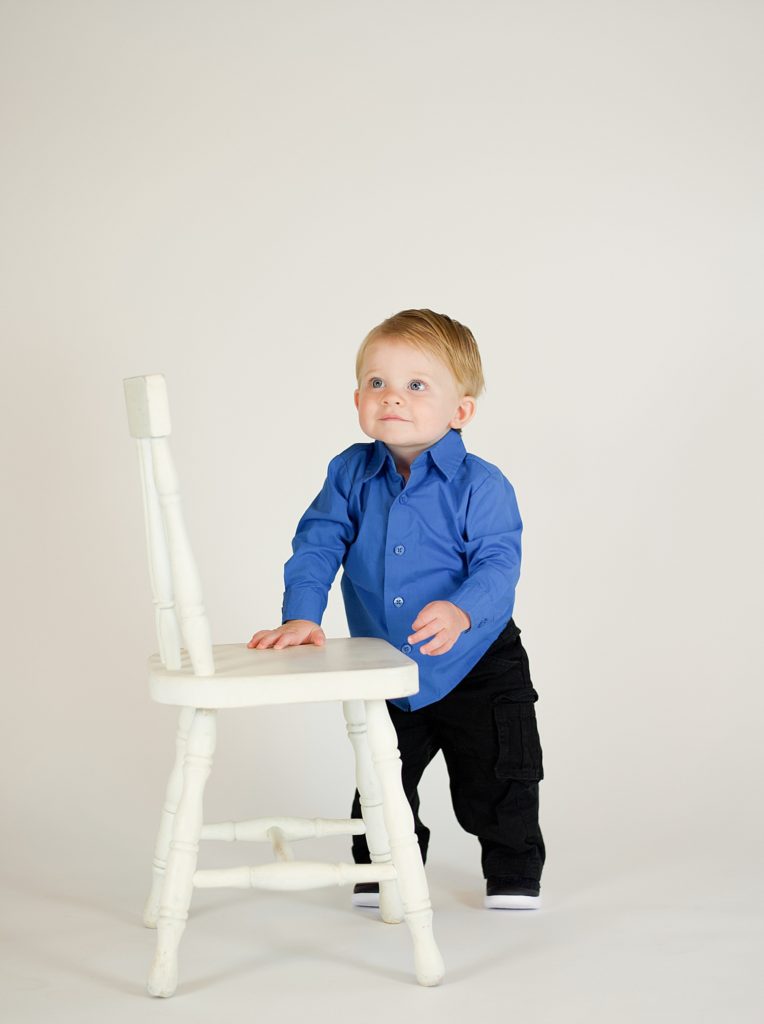 toddler in blue button up shirts by white chair during first birthday cake smash