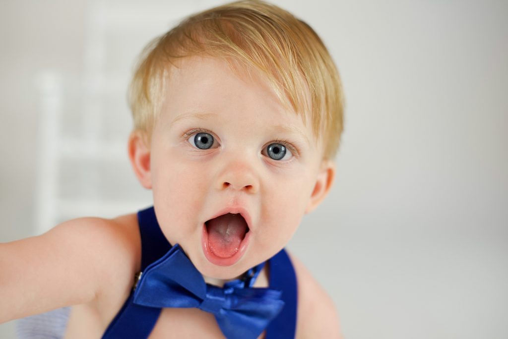 toddler in suspenders with blue bow tie plays during first birthday cake smash