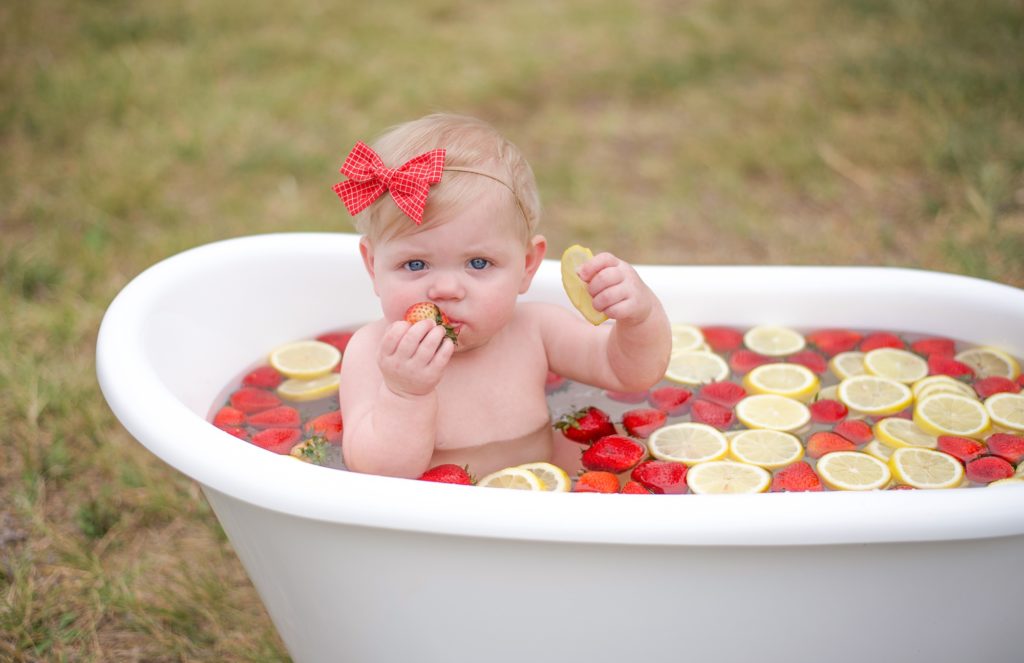 9 month old in red bow sits in tub with strawberries and lemons for milestone photos 