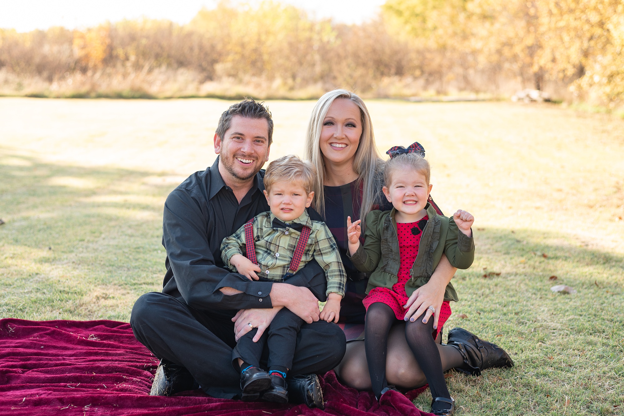 family in red, black, and green holiday outfits sit on red blanket during Little Elm backyard family portraits