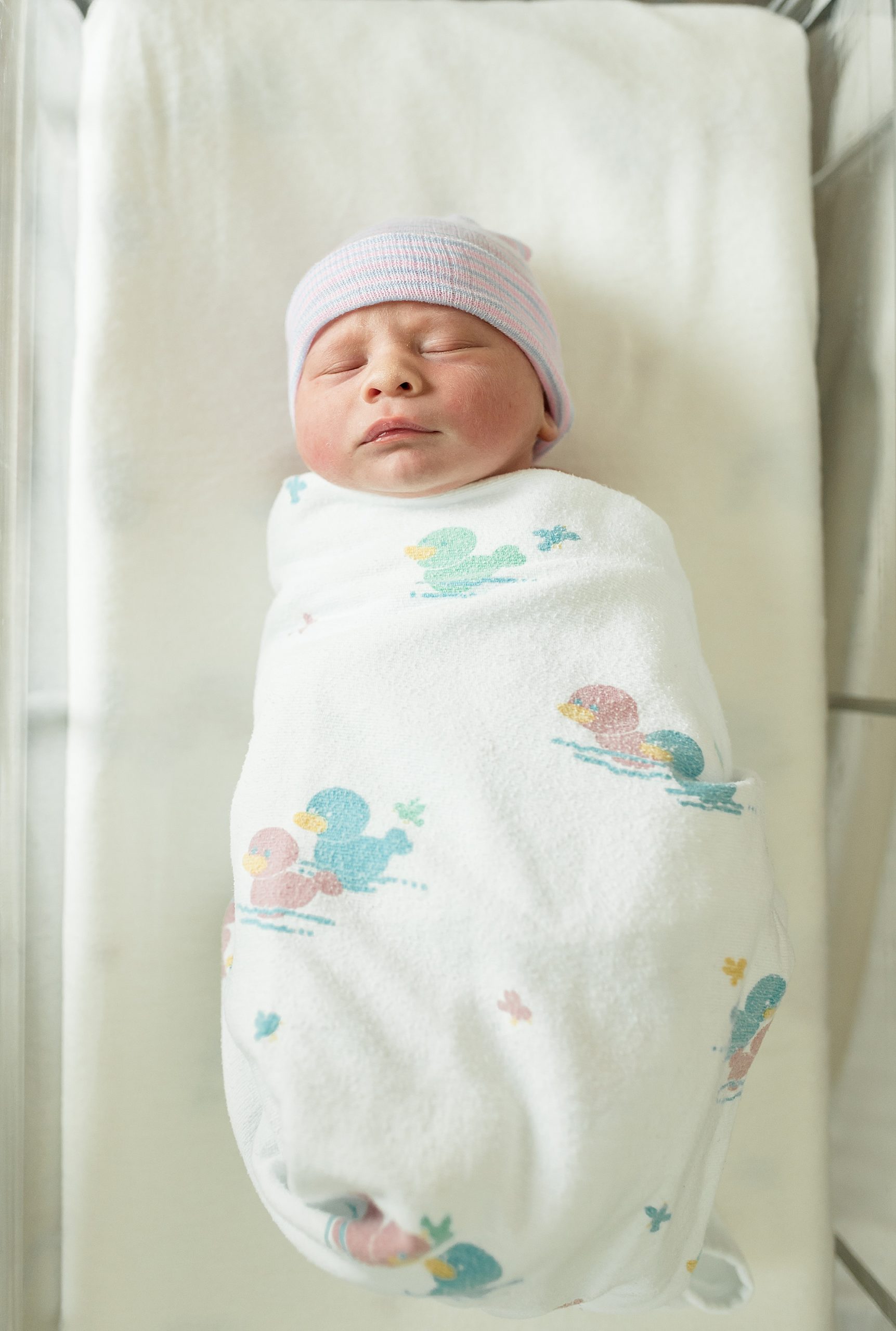 newborn lays in crib in white swaddle and pastel hat 