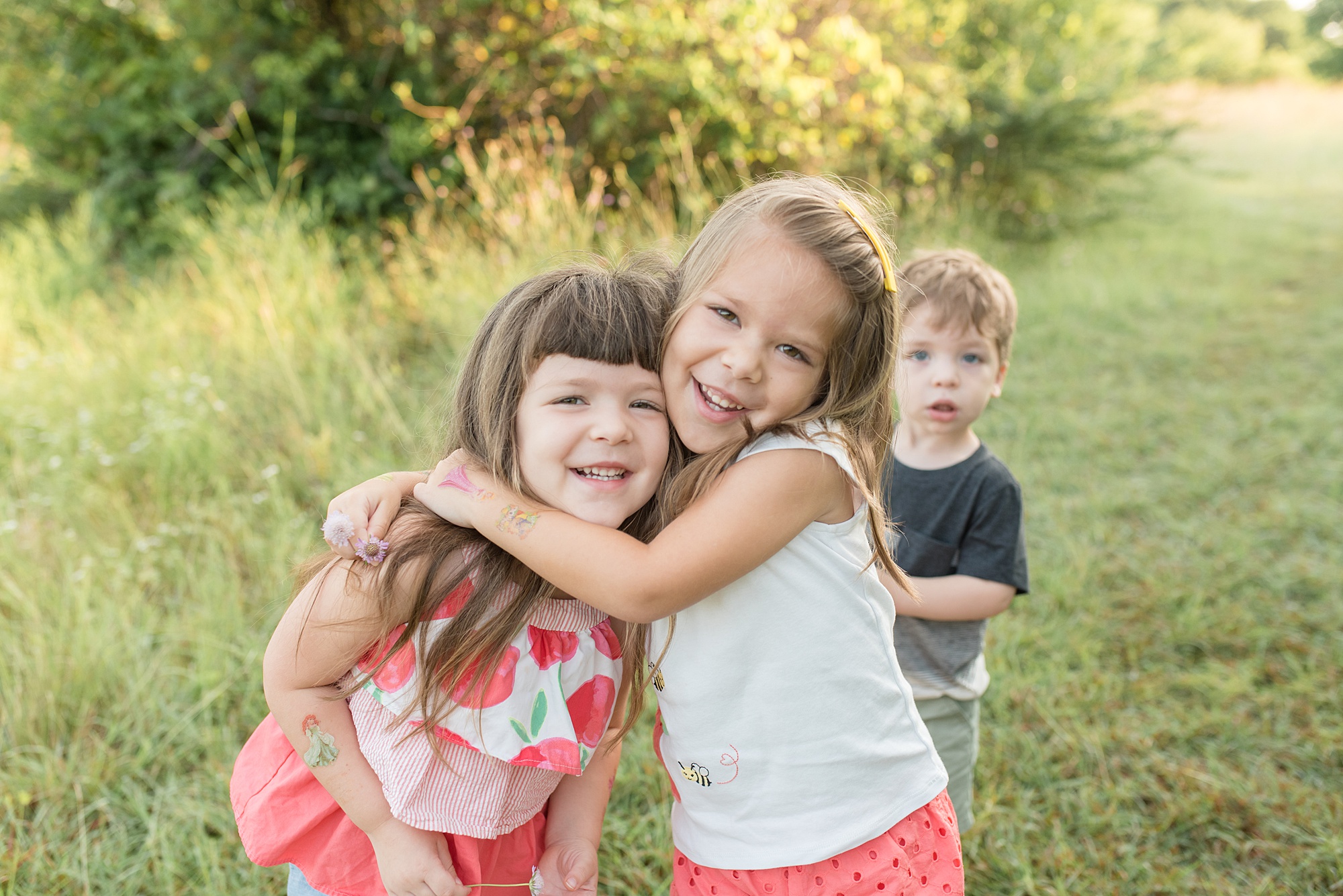 sisters hug with brother in background in Frisco Texas photographed by Lindsey Dutton Photography