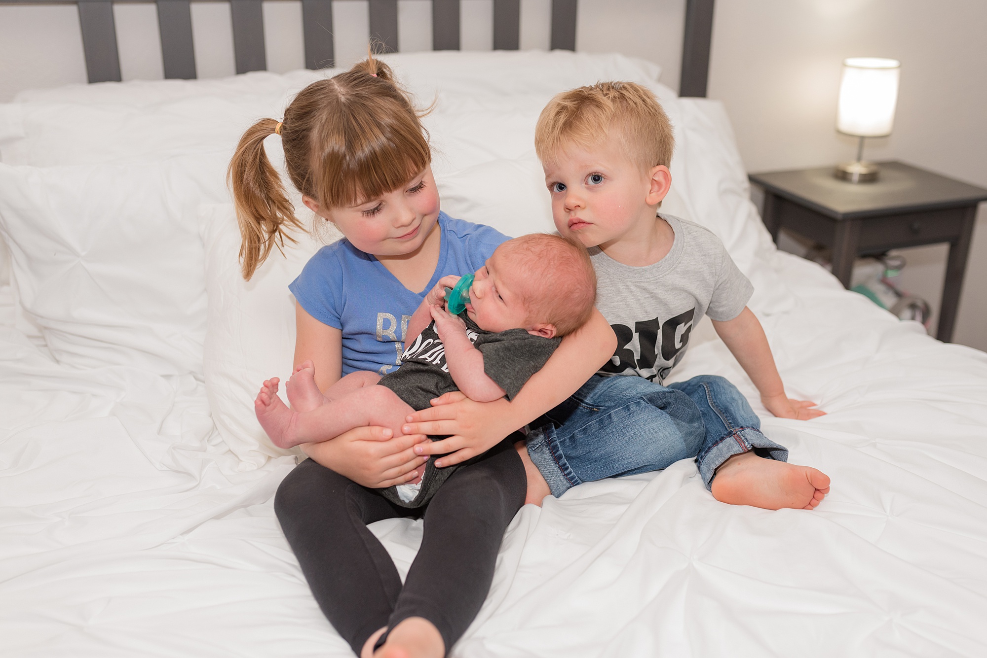 older sister holds newborn brother with the middle brother nearby on the bed 