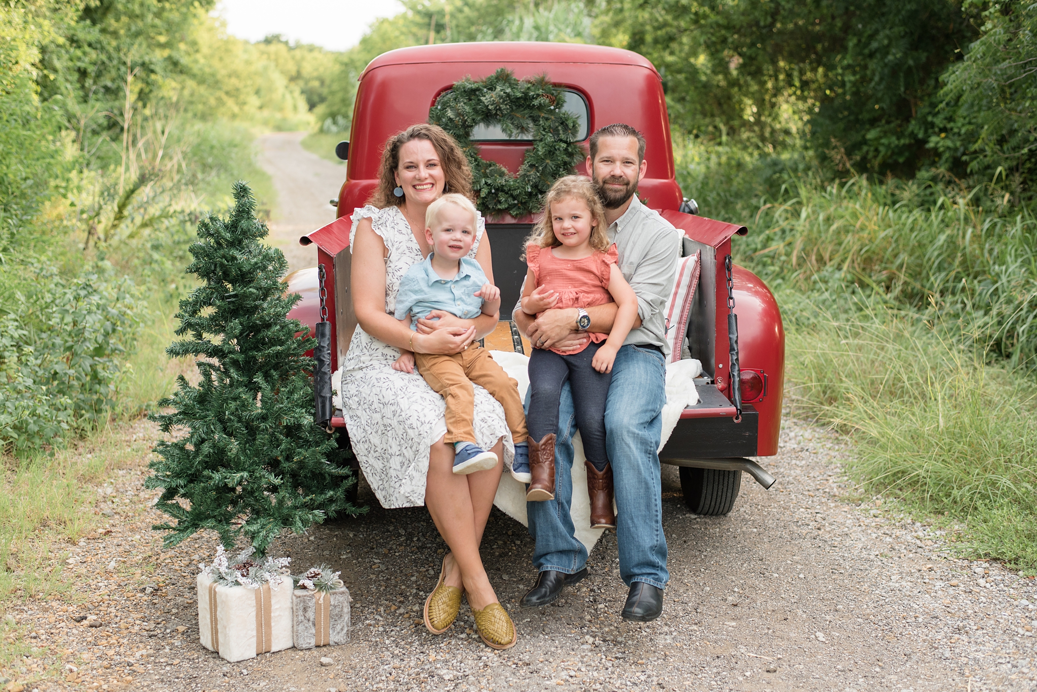 family sits in the back of a red pick up truck with a green pine tree during family photo session