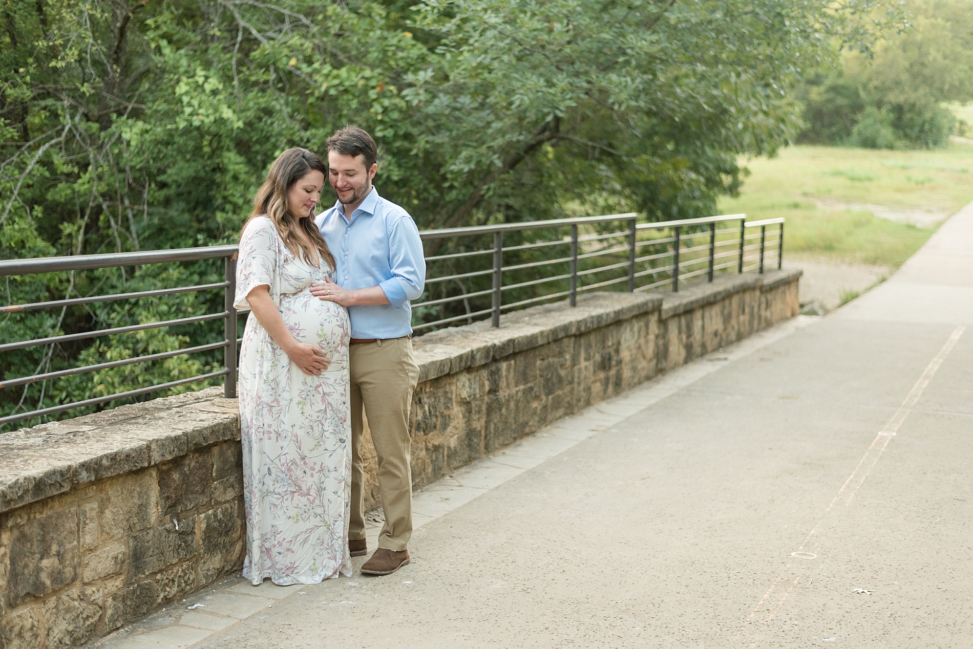 couple looks down at pregnant belly standing on bridge in Plano, TX