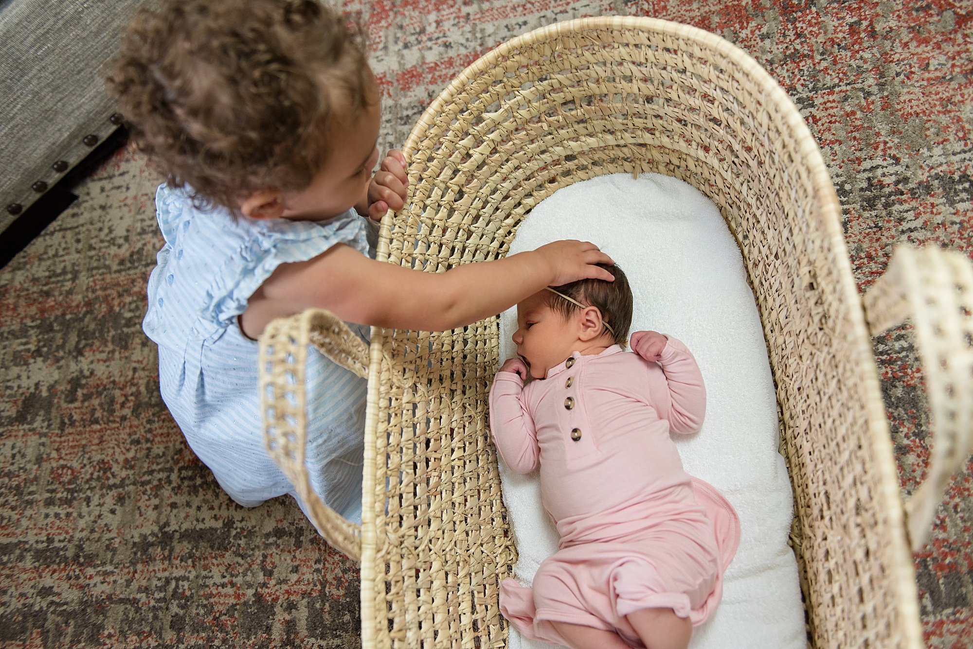baby girl in pink onesie in woven basket with big sister watching over