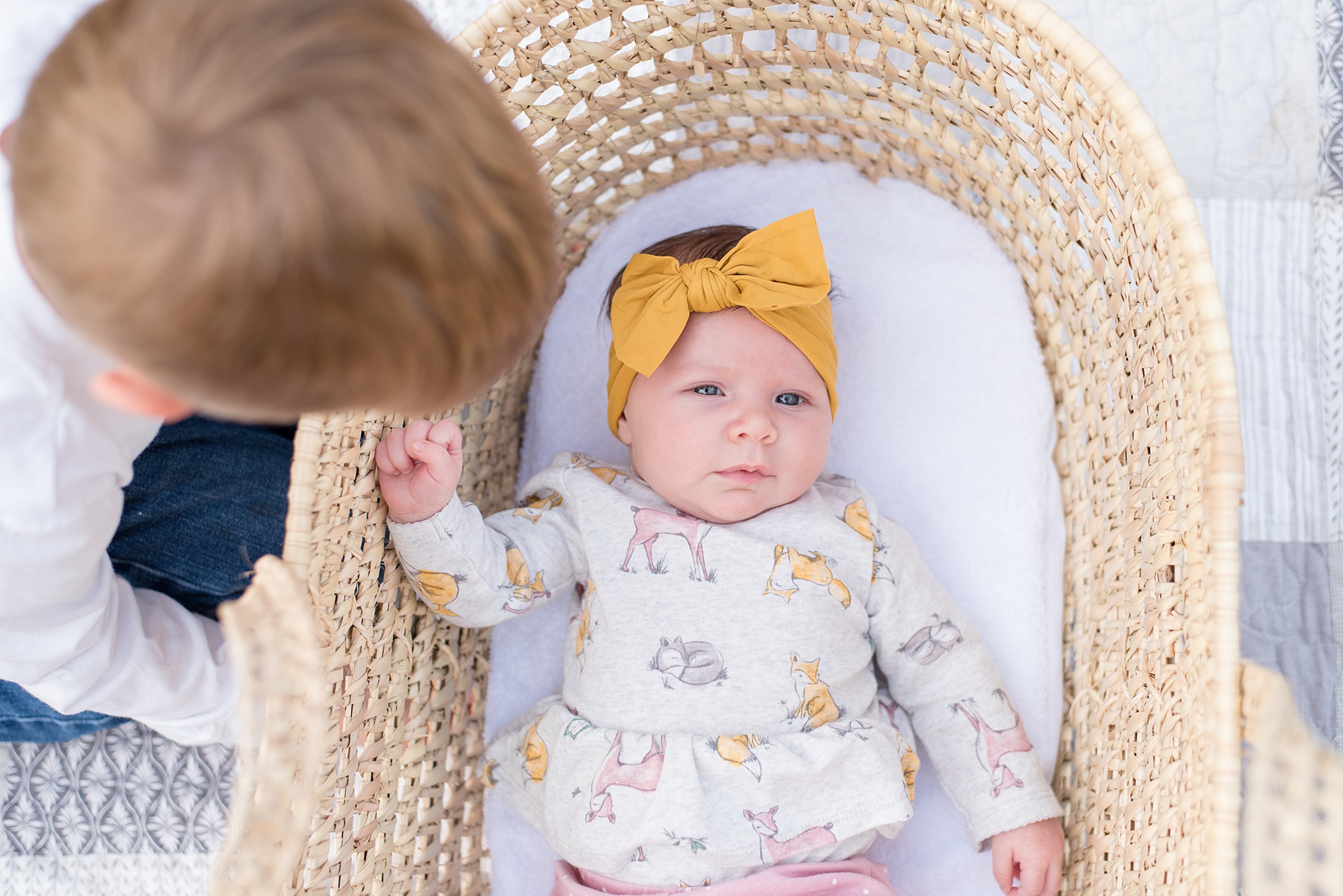 baby girl in yellow headband lays in bassinet while big brother checks in on her