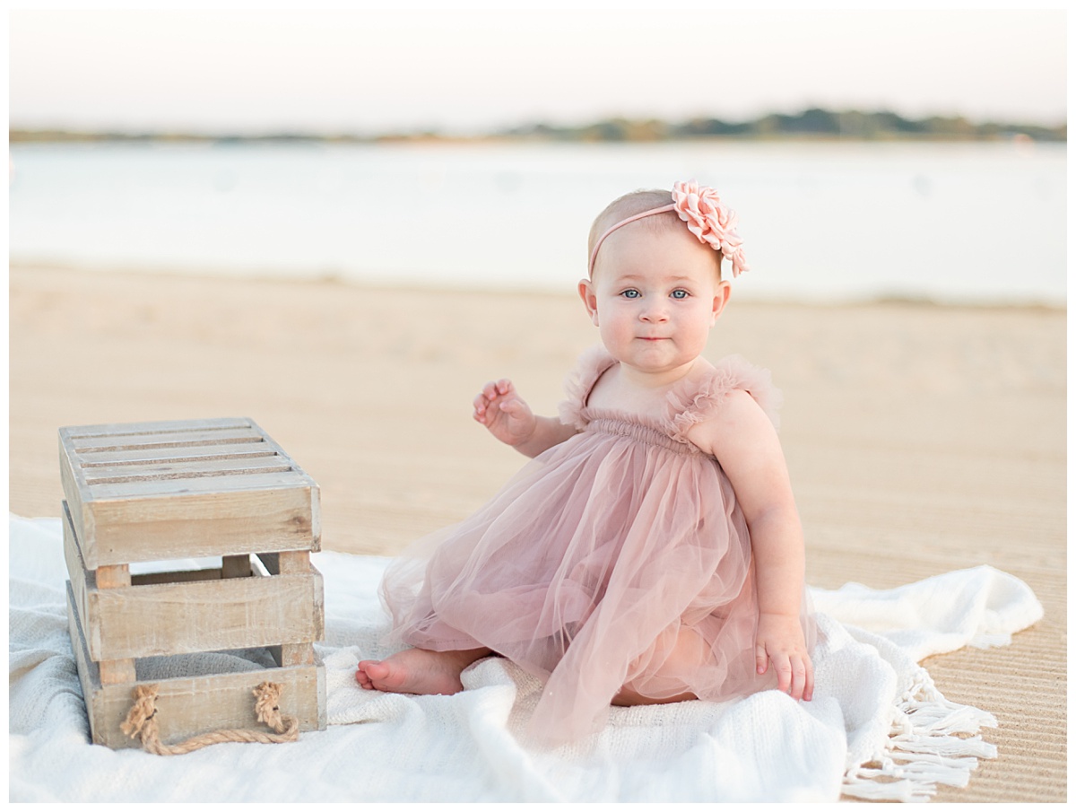 Baby girl in pink, tulle dress sitting on the beach for her cake smash session in North Dallas taken by Lindsey Dutton Photography, a Dallas Cake Smash Photographer.
