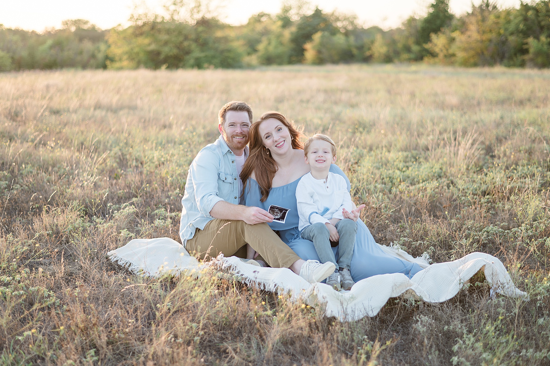 Family sitting in field holding sonogram photographed by Lindsey Dutton Photography, a Dallas maternity photographer
