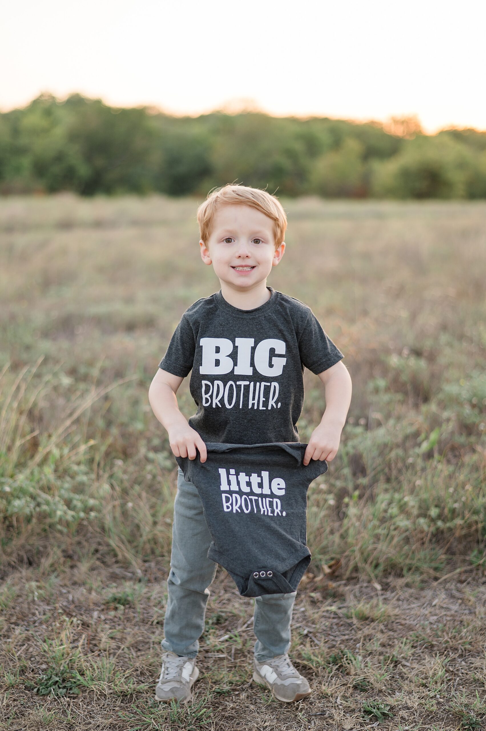 boy in big brother shirt holds up little brother onesie during pregnancy announcement photoshoot taken by Lindsey Dutton Photography, a Dallas maternity photographer
