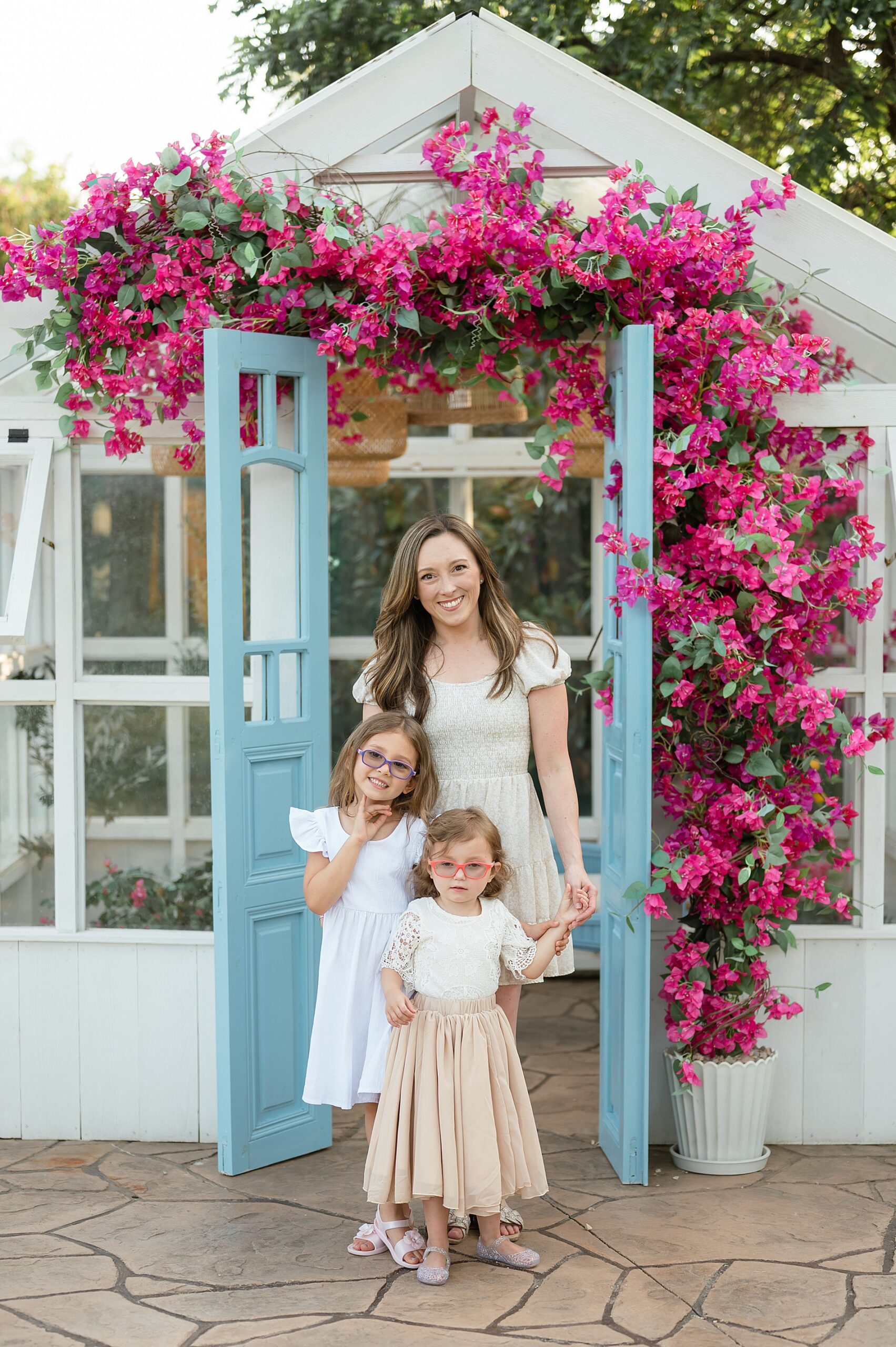 mom with two daughters stand in front of greenhouse decorated in pink flowers photographed by Lindsey Dutton Photography, a Dallas Family photographer
