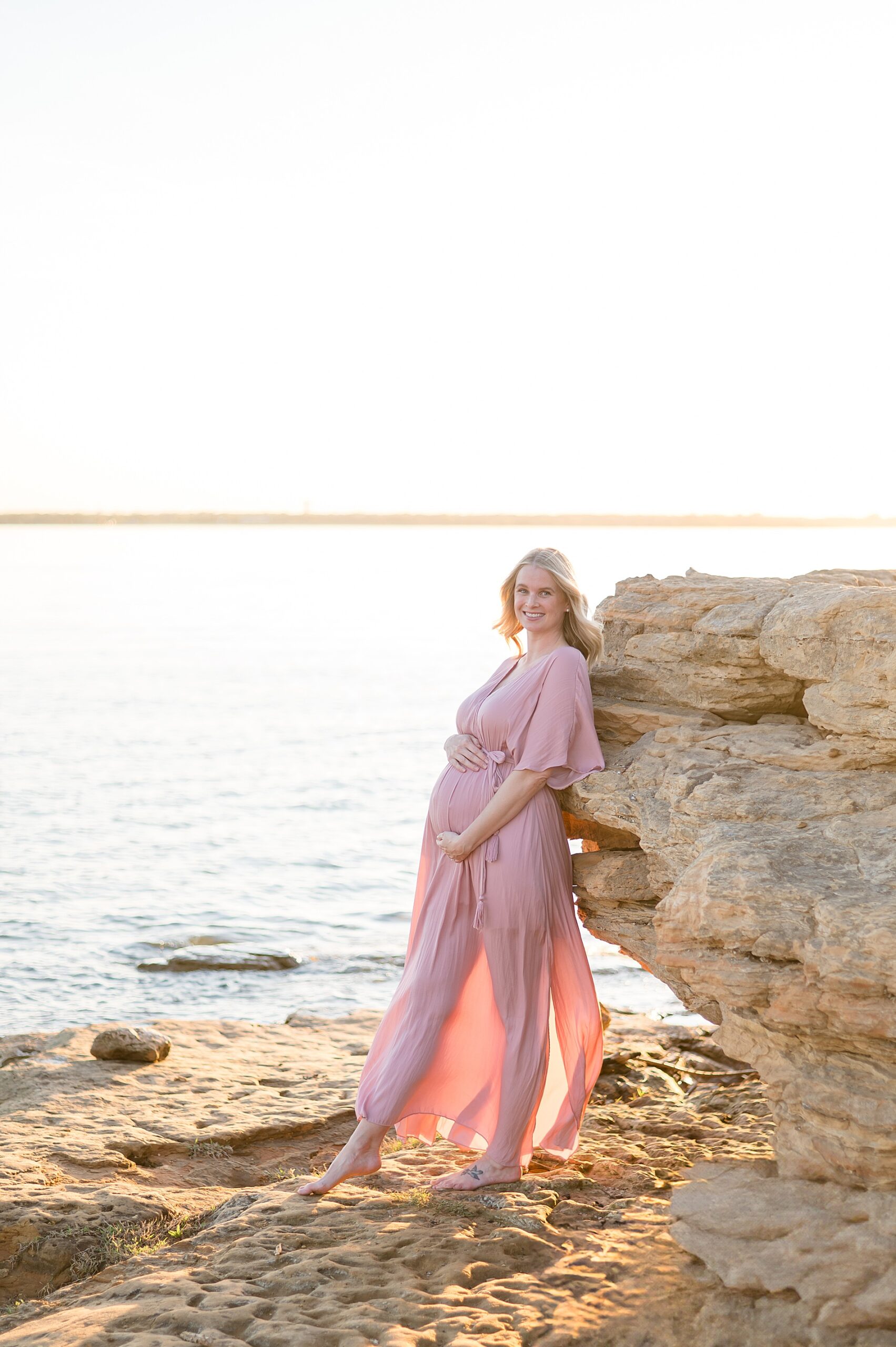 expectant mom in flowy pink dress leans against large rock from Winter Lakeside Maternity Session in Flower Mound, TX