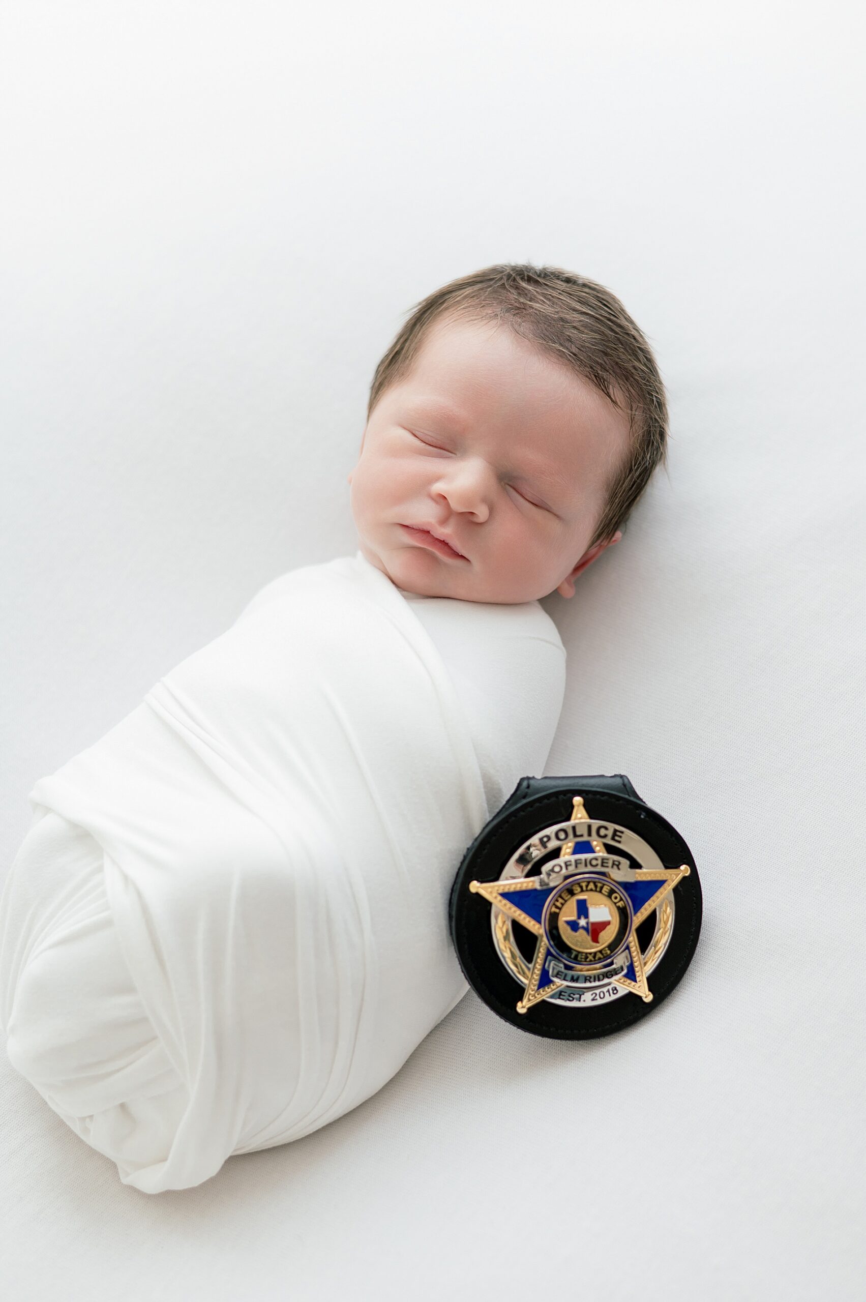 newborn swaddled in white blanket with police badge next him