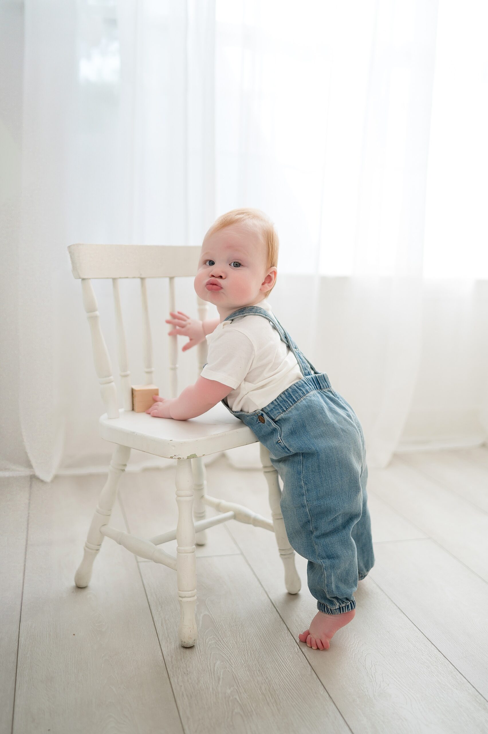 little boy in jean overalls makes adorable faces during one year milestone session 