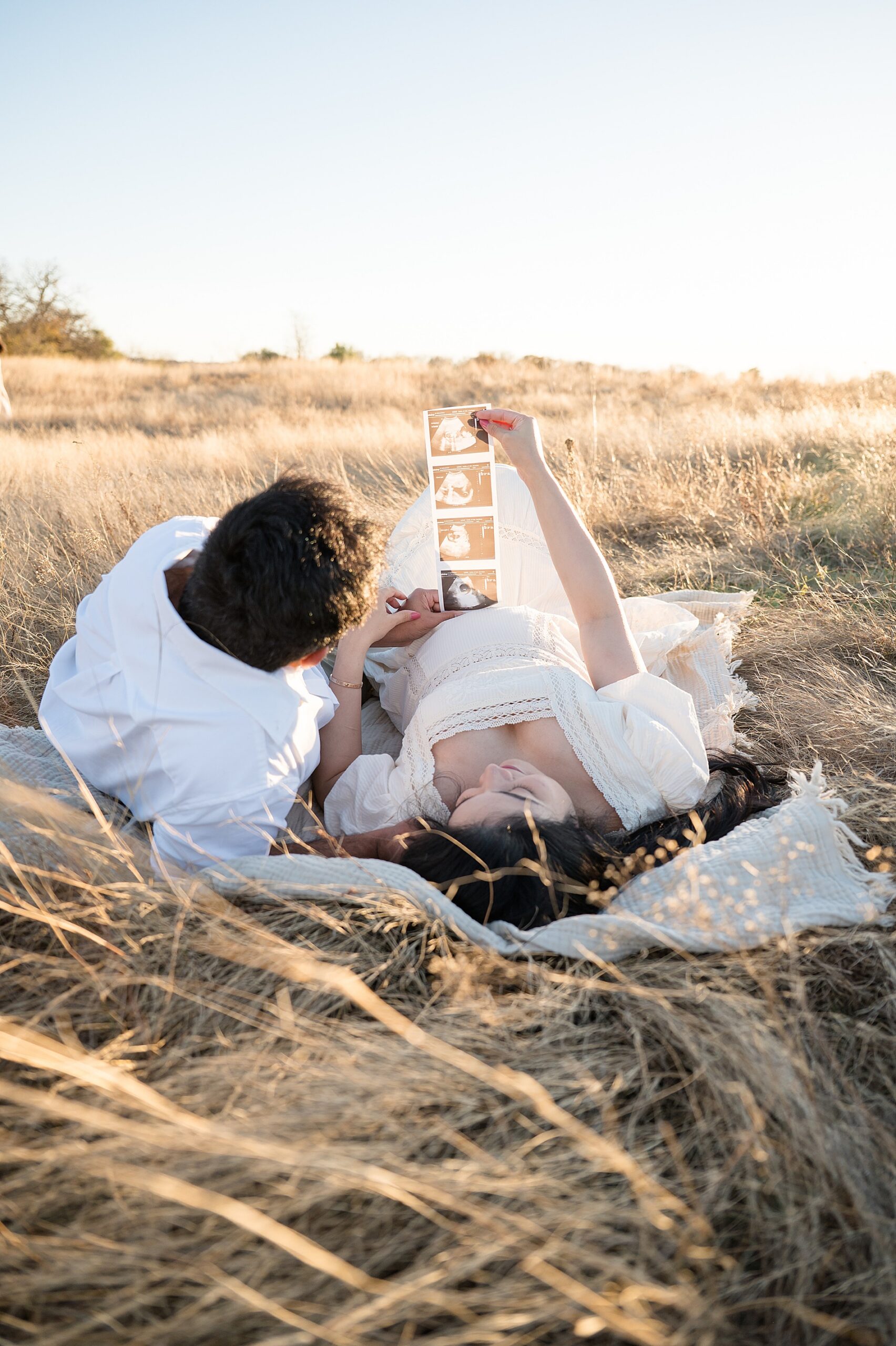 couple lay in grassy field together looking at ultrasound photo during Flower Mound, TX Maternity session 